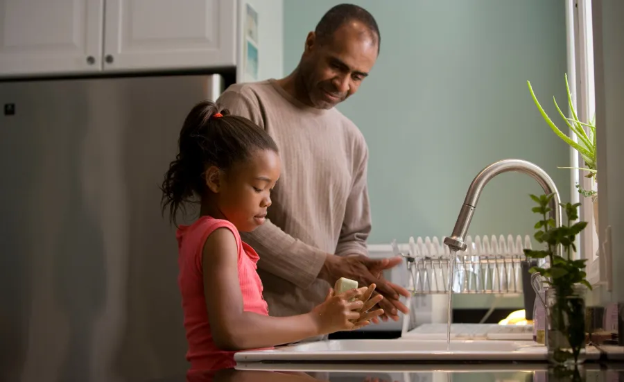 A father smiling down at his young daughter as she washes her hands carefully with soap.