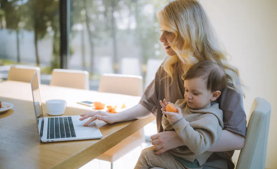 A woman sitting at a table using a laptop with one hand and a child sitting on her knee eating an orange 