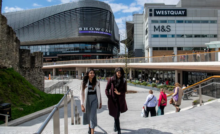 Two students talk whilst walking across a large paved area with several large shops.