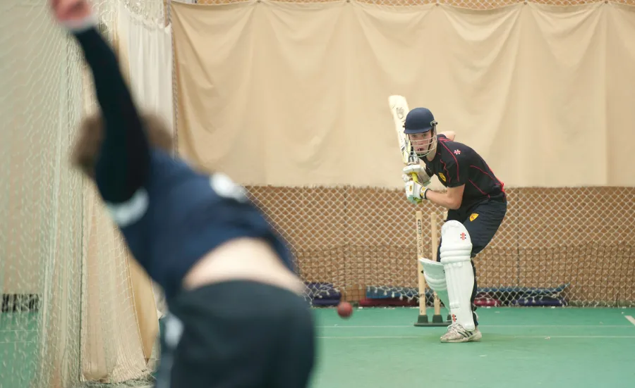 Two students in cricket nets. One, in protective gear, focuses intently on hitting a fast moving ball bowled by the other.
