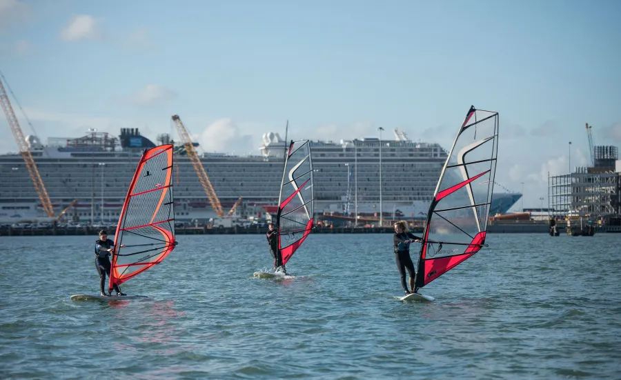 Three people windsurfing beneath blue skies on a sunny day. A large cruise ship is visible behind them, docked in port.