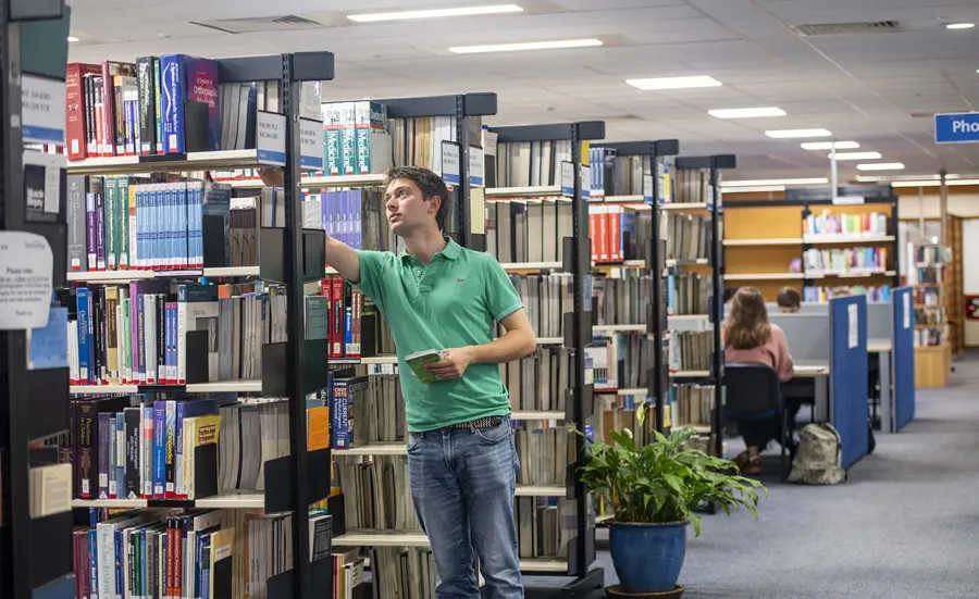 A student selects a book from a shelf while other students work at desks in the background