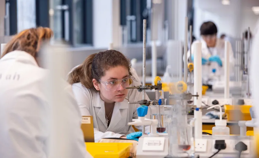 A student in a lab coat crouches down to closely monitor a chemistry experiment being conducted in a large piece of glassware.