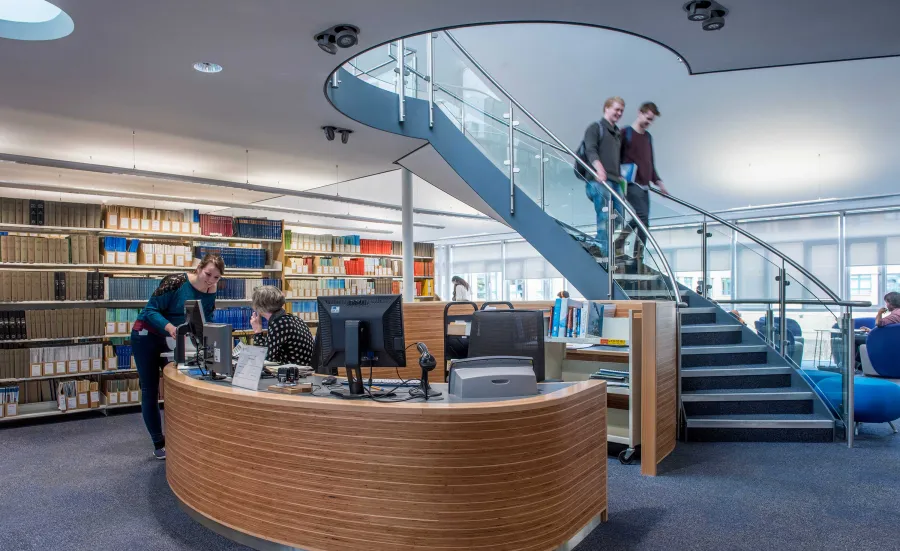 A bright, modern library reception area with shelves full of books. A lady at the desk helps someone. Two students descend a spiral staircase from an upper floor.