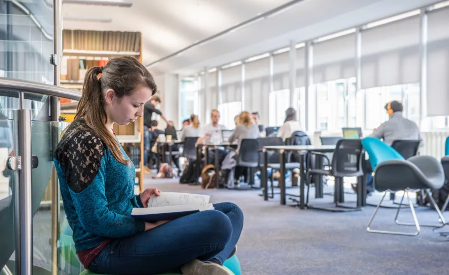 A student sits cross-legged reading a book in a bright, airy library. Behind her are shelves of books and other students working at desks.