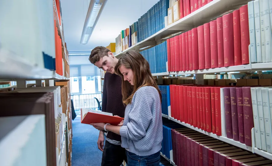 Two students in a narrow library aisle, surrounded by shelves of books on both sides. They are reading a book pulled from one of the shelves.