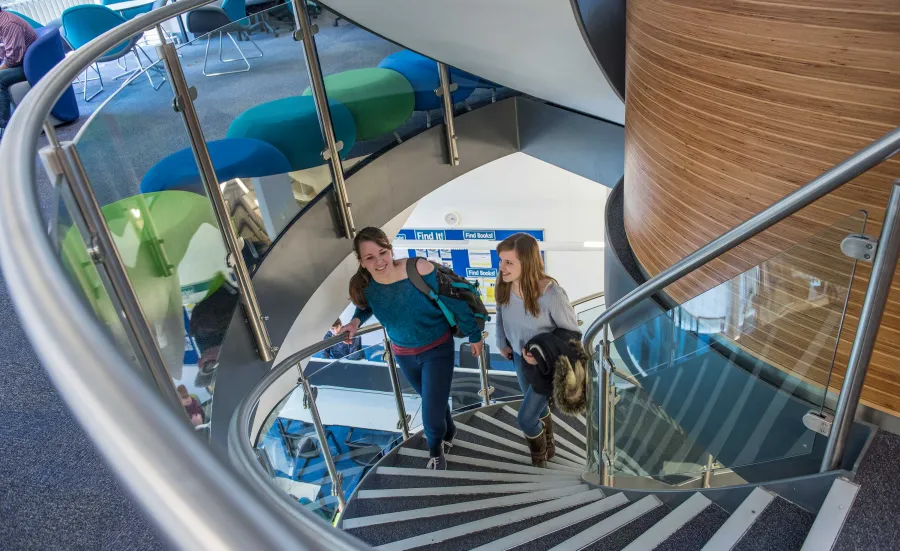 Two students ascend a set of spiral stairs from a lower to an upper floor. Study spaces, desks and stools are visible on the upper floor.