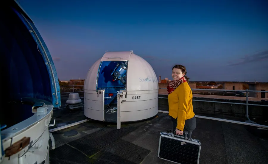 A student stands on the roof of a building at night time. She smiles towards the camera. On the roof are two small dome-shaped observatories containing telescopes.