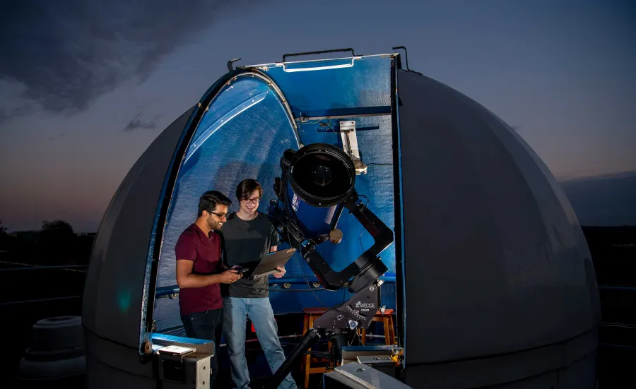 A small, domed rooftop observatory opens onto a starry night sky. Inside the observatory, two students use a telescope and a laptop to collect data.