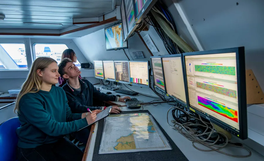 Two students sit at a desk inside a room on a boat. They observe a bank of computer screens displaying various data and information.