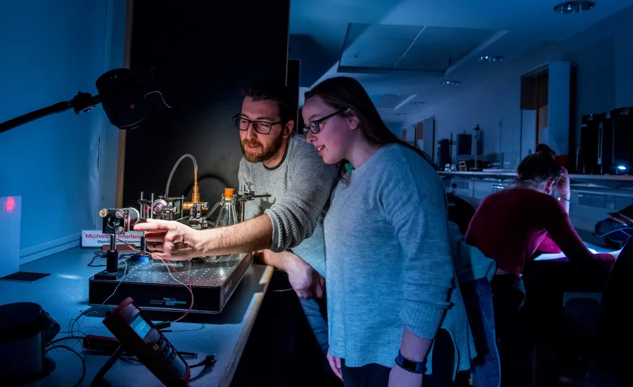 Two students set up a small experiment with a laser, in a darkened laboratory, surrounded by technical equipment.