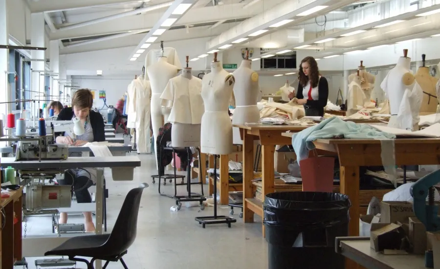 A wide shot of the fashion studio, full of students hard at work. To the right, a row of large workbenches are mixed in with over a dozen mannequins wearing various designs. To the left, a row of sewing machines.