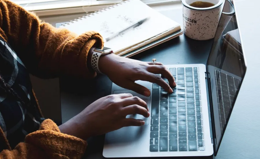 Fingers typing on a laptop with notepad, pen and mug in the background