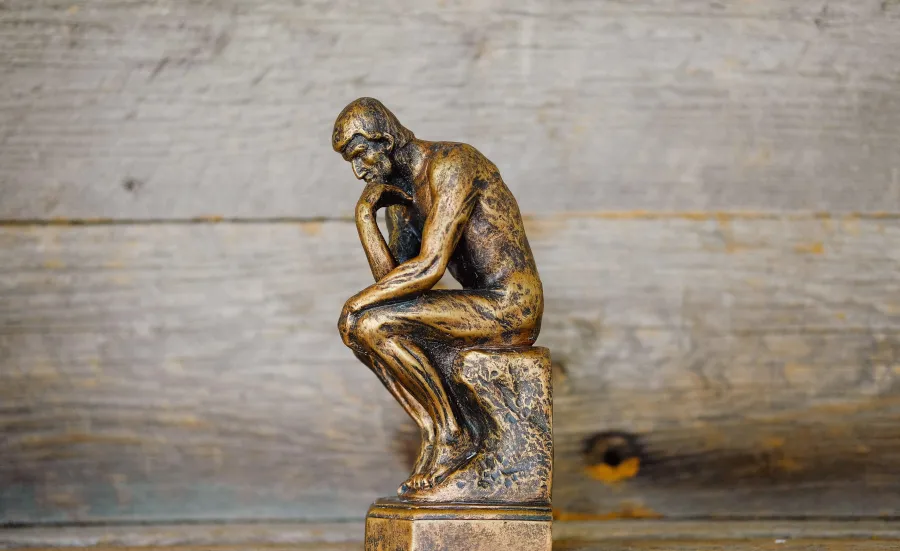 A small replica of 'The Thinker' - a bronze sculpture by Auguste Rodin depicting a man hunched over on a rock in contemplation - set against a backdrop of wood panels.