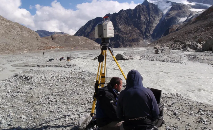 Two researchers sitting together on the ground, surveying data, in a mountain landscape.