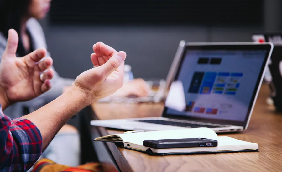 Laptop on a table with notepad and phone, user's hands in foreground 