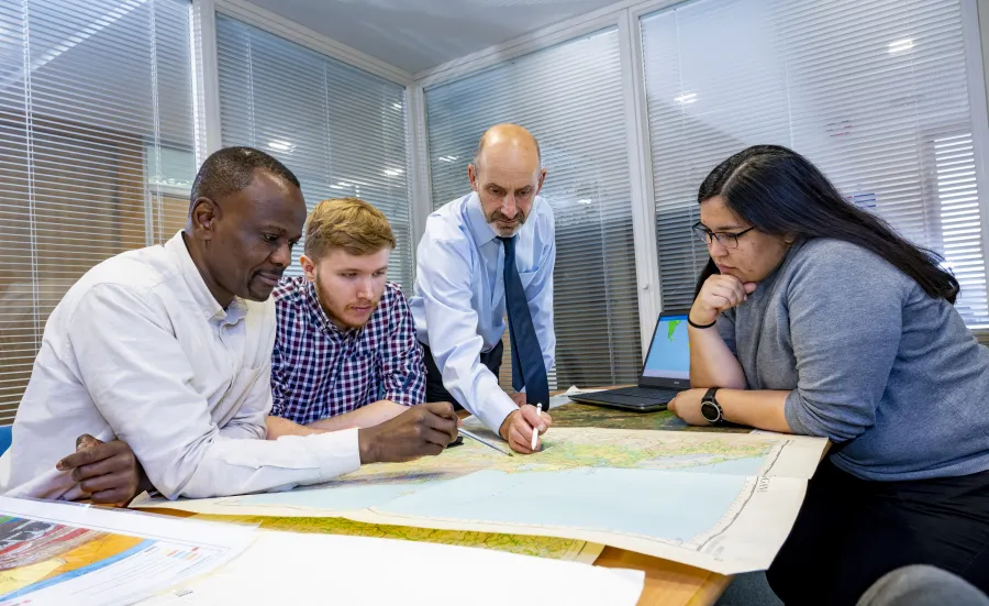 A Southampton Geospatial researcher talks with team members, standing over a map, with a laptop with a data visualisation to the side