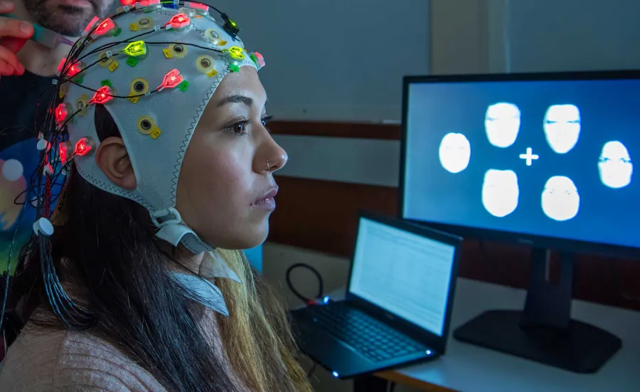 A study participant wears an EEG cap and is seated close to a screen displaying data.
