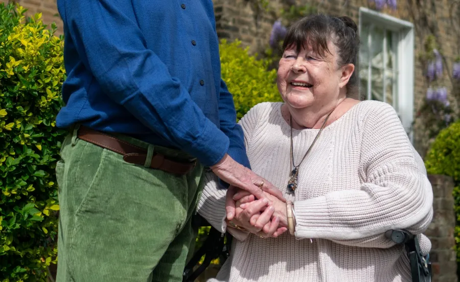 A woman in late middle age sits and holds hands of a man standing next to her