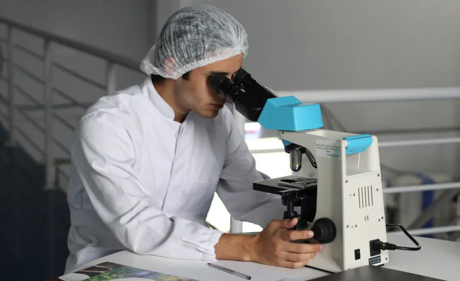 A researcher in a white lab coat looks through a microscope.