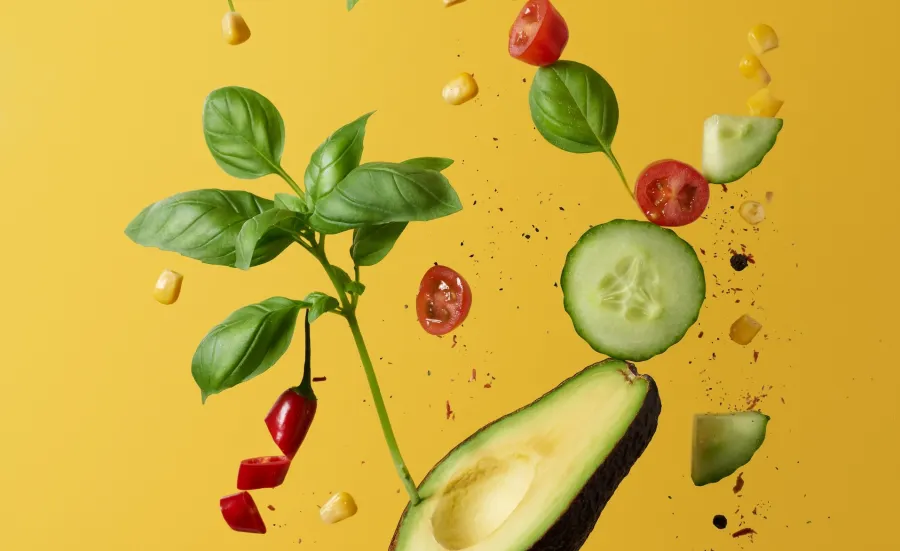 Half an avocado, halved cherry tomatoes, a red sliced chili pepper, basil leaves, cucumber slices and fine herbs fly into the air against a yellow background