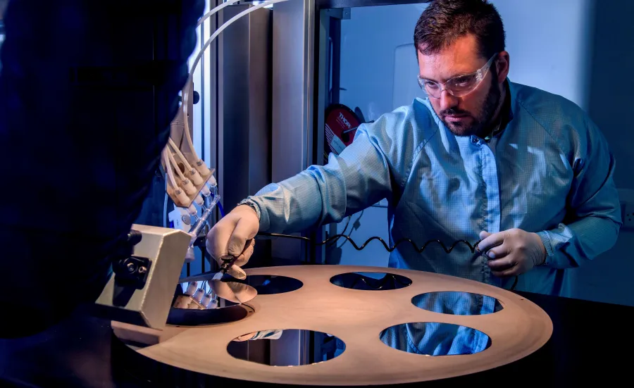 A bearded young scientist in protective overalls and glasses works with a flame hydrolysis deposition (FHD) system