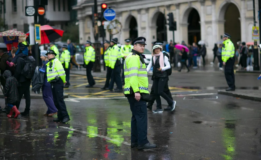 A number of police officers monitor crowds of people on a rainy London street. They are wearing high visibility yellow jackets.