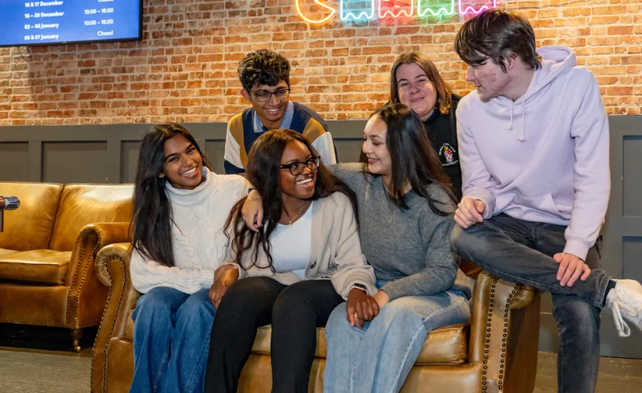 A group of six students sat on or around a brown leather sofa in a students' union. All of the students are smiling, and some are holding hands or have their arms around each other. There is a neon sign of video game characters and a TV mounted on the wall behind them.