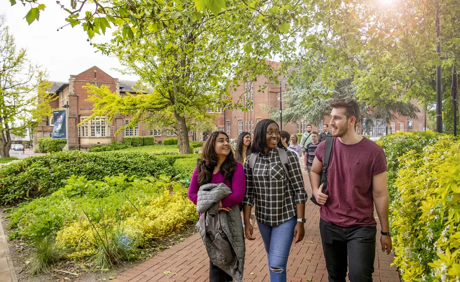 Students with backpacks walk together across Highfield campus with the Hartley Library in the background. Trees are in leaf and sunlight is visible through the foliage.