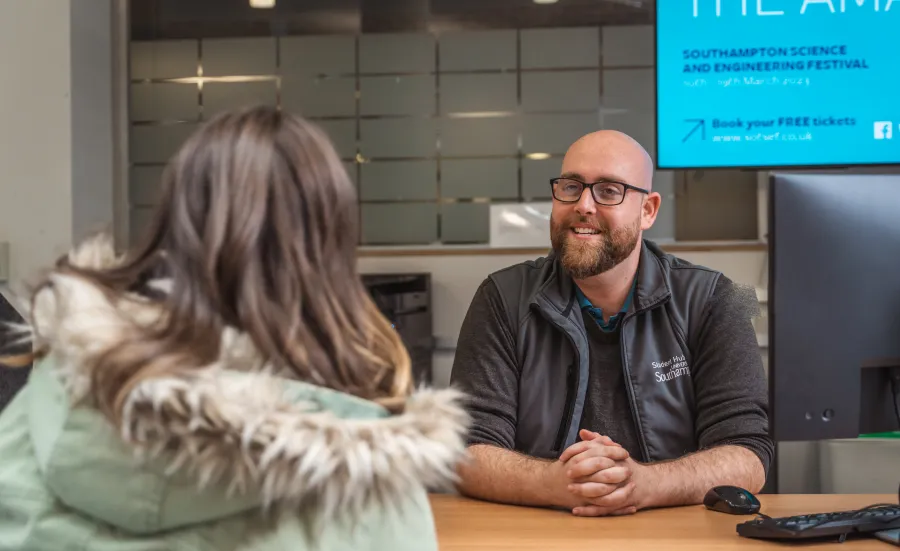 A University of Southampton staff member sits behind a desk while speaking with a student. The staff member is smiling and wearing University of Southampton-branded clothing.