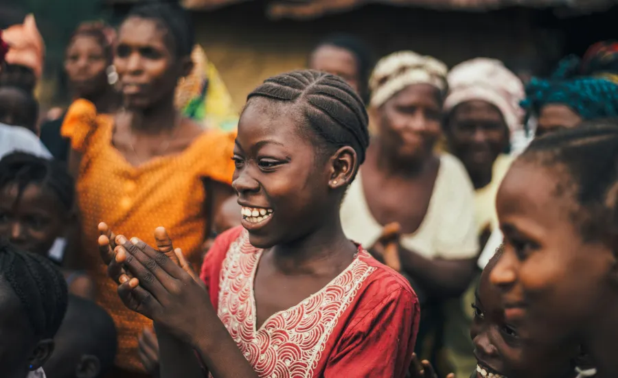 A young girl with braided hair claps and smiles amidst a group of villagers outdoors, capturing the vibrant spirit of Sierra Leone.