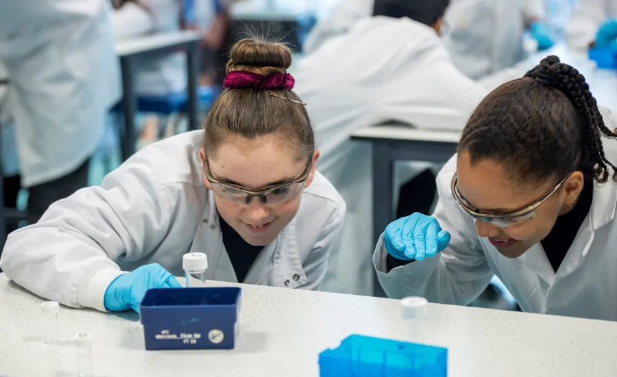 Two school children in lab coats and safety goggles examining laboratory equipment with focus, surrounded by others in a laboratory setting.