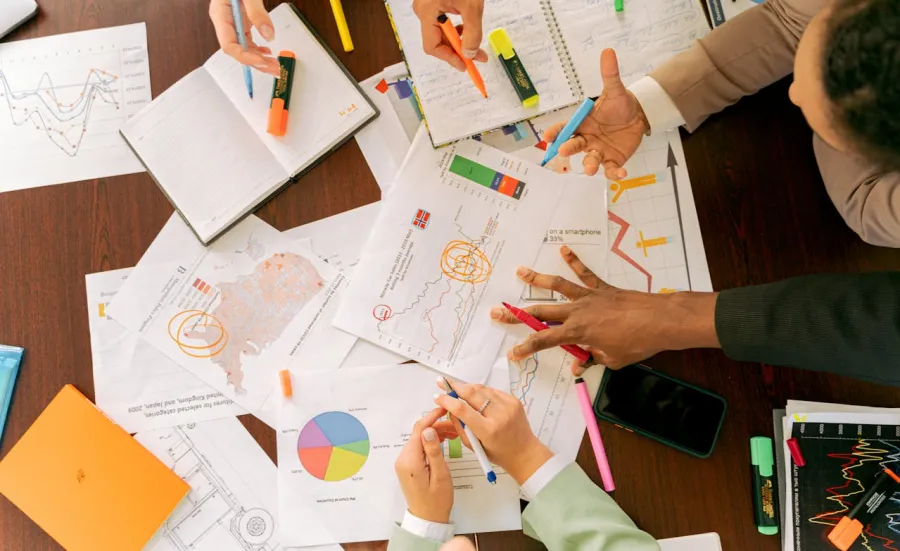 Looking down at a work surface covered in paper charts and notebooks. There are hands holding coloured pens on the paper documents.