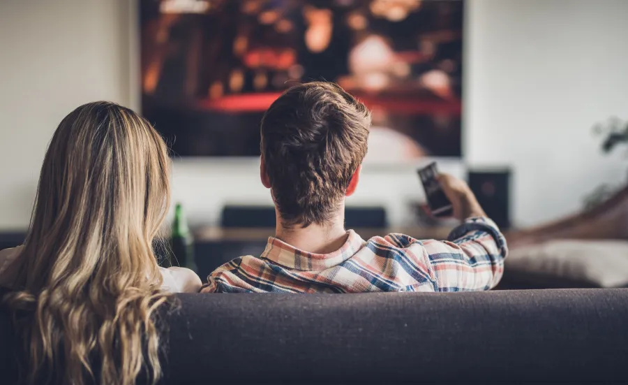 A couple sitting on a sofa, watching a wall mounted TV. The man is pointing the remote at the screen.