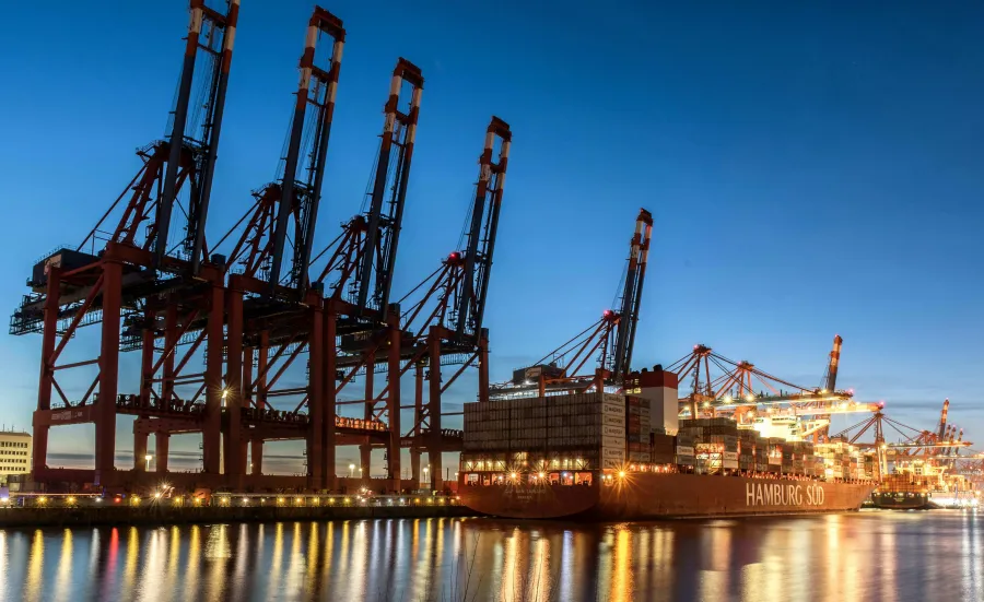 Large fully loaded container ships docked at port at dusk. Large cranes are unloading the cargo. The water has reflections of the lights