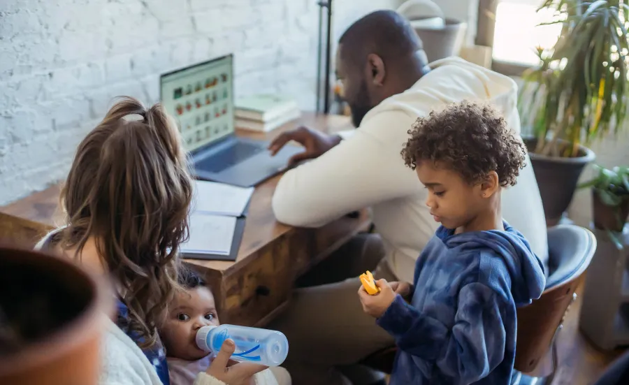 Family at home with father working on laptop, mother feeding baby, and young child playing nearby.