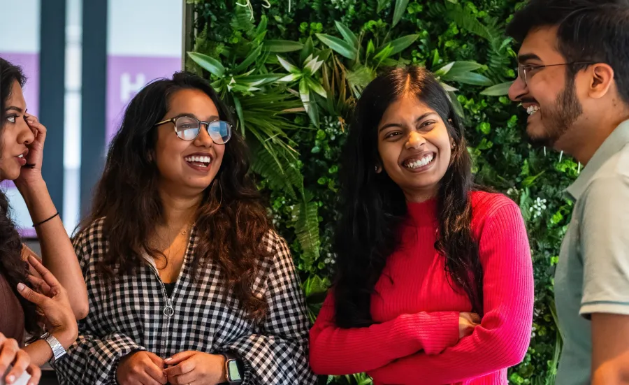 A group of smiling students having a conversation in front of a vibrant green wall with plants.