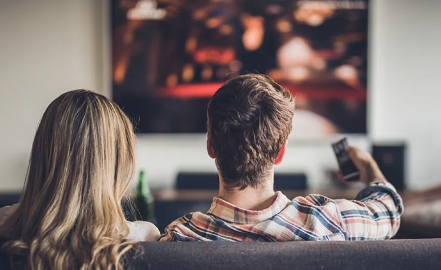 2 people sitting on a soda watching a large wall mounted TV. The man is pointing the remote at the screen.