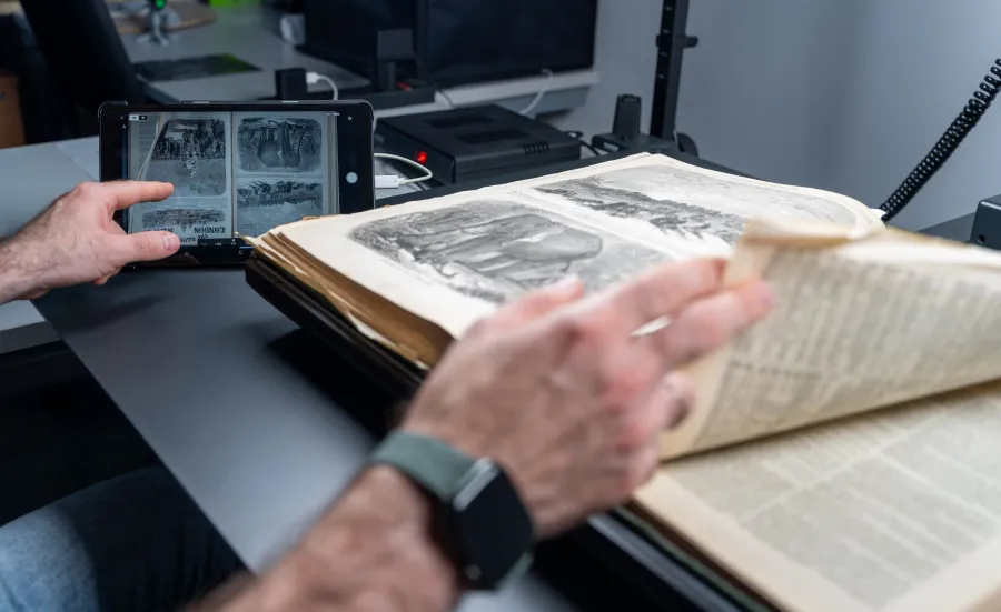 A close-up of a person's hands working with an open historical book on a specialized scanning table. One hand is turning a delicate page with illustrations, while the other holds a tablet displaying a digital scan of the same page. The workspace includes modern scanning equipment and cables in the background, blending traditional and digital technologies.