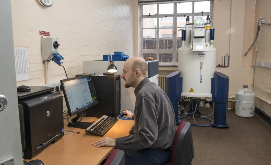 A man working  on a computer in a room with a Nuclear Magnetic Resonance machine in the background