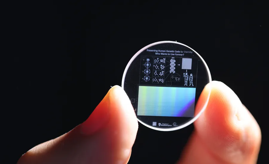 Zoomed in view of a hand holding a 5D memory crystal between two fingers. The memory crystal is a small clear disc with diagrams of human DNA on the surface.