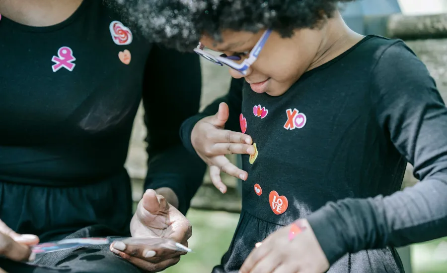 A young child is choosing stickers from a sheet held by an adult. The child is adding the colourful stickers to their top.  