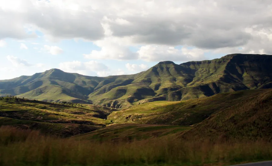 A landscape of rolling hills with long grass in the Lesotho Highlands. There is a mountain range on the horizon, and the sky is mainly cloudy with some clear blue areas.