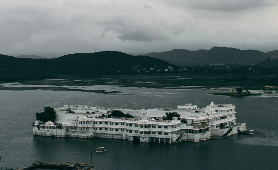 The Taj Lake Palace in Udaipur, Rajasthan, India. It is a rainy day and there are dark clouds surrounding the hills in the distance.