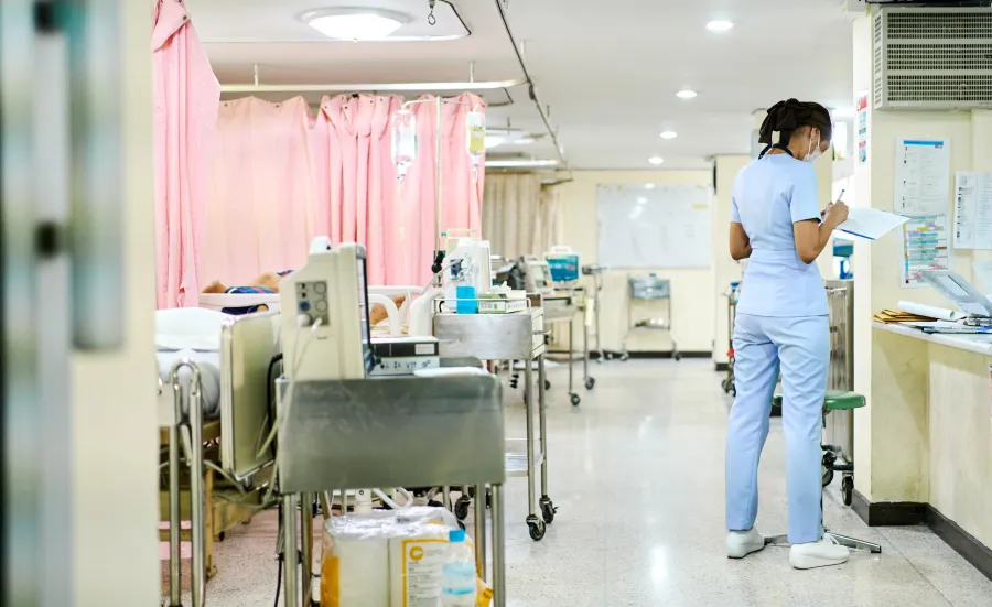 A hospital ward showing a trolley of equipment at the end of each bed. There is 1 member of staff facing away from the camera completing a chart.