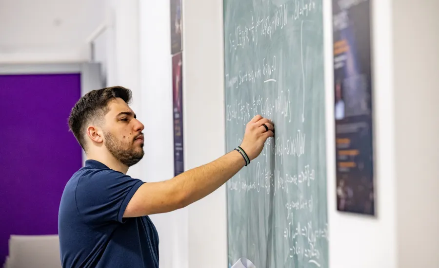 A student writing a mathematical equation on a blackboard, in a study and social space on a university campus.