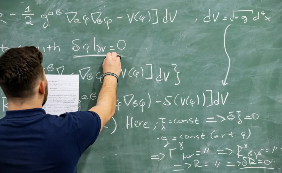 A student writing a mathematical equation on a blackboard, in a study and social space on a university campus.