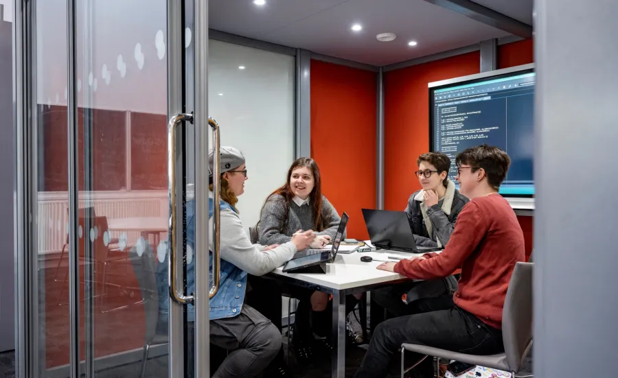 Four people sat around a table in a small meeting room. Two of the people have laptops open on the table, and there is a TV screen mounted on the wall showing code. The walls of the meeting room are red, and the glass door to the room is open.