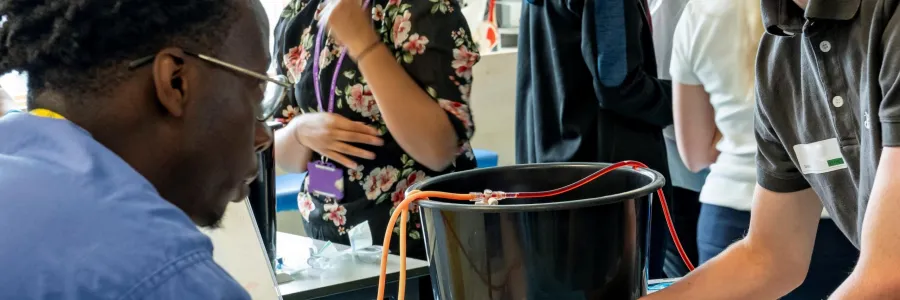 Two people practice a medical procedure on a mannequin's arm while others observe or converse in a well-lit clinical education setting.