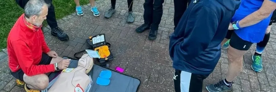 A group of people is observing a CPR training demonstration outdoors, with one person performing the demonstration on a training mannequin next to an AED device, in a sunny, grassy area.